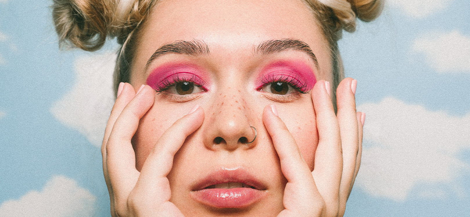 Close up of woman's face with hair in two buns, pink eyeshadow on eyelids, and chin in hands